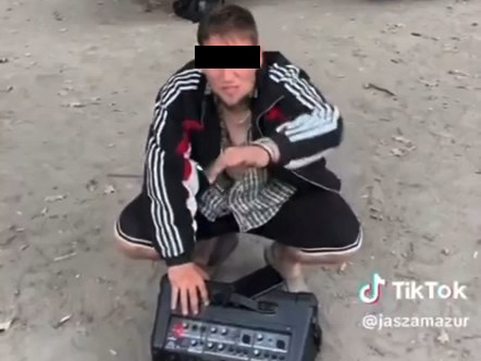 Mężczyzna w sportowej kurtce klęczy na ziemi, obsługując urządzenie audio w leśnym otoczeniu.