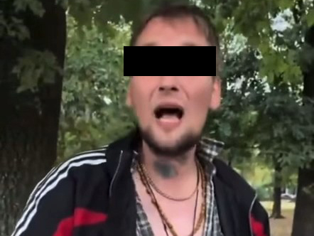 Mężczyzna w dresie z tatuażem na szyi, głośno przemawiający w parku, otoczony drzewami.