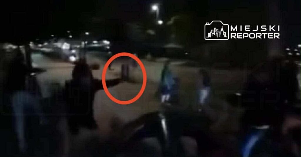 Grupa osób zebrana nocą na ulicy, jedna osoba trzyma rękę uniesioną w geście. W tle widać zaparkowane samochody.