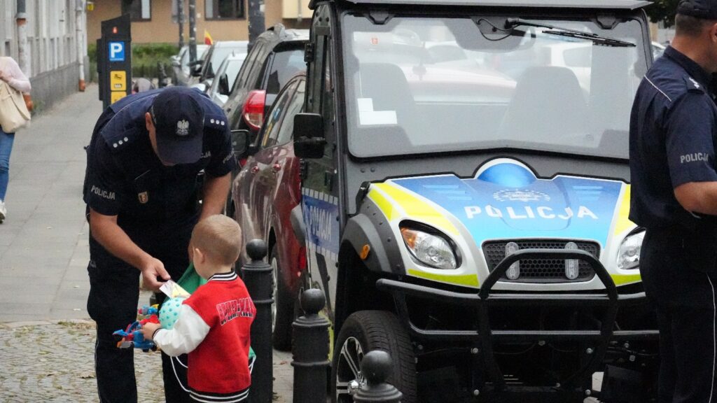 Policjant wręcza zabawkę chłopcu w czerwonej kurtce na ulicy, obok radiowozu z oznaczeniem "POLICJA".