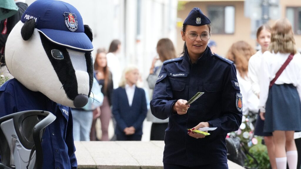 Policjantka w mundurze rozdaje ulotki obok maskotki w kształcie borsuka na ulicy, w tle grupka dzieci.
