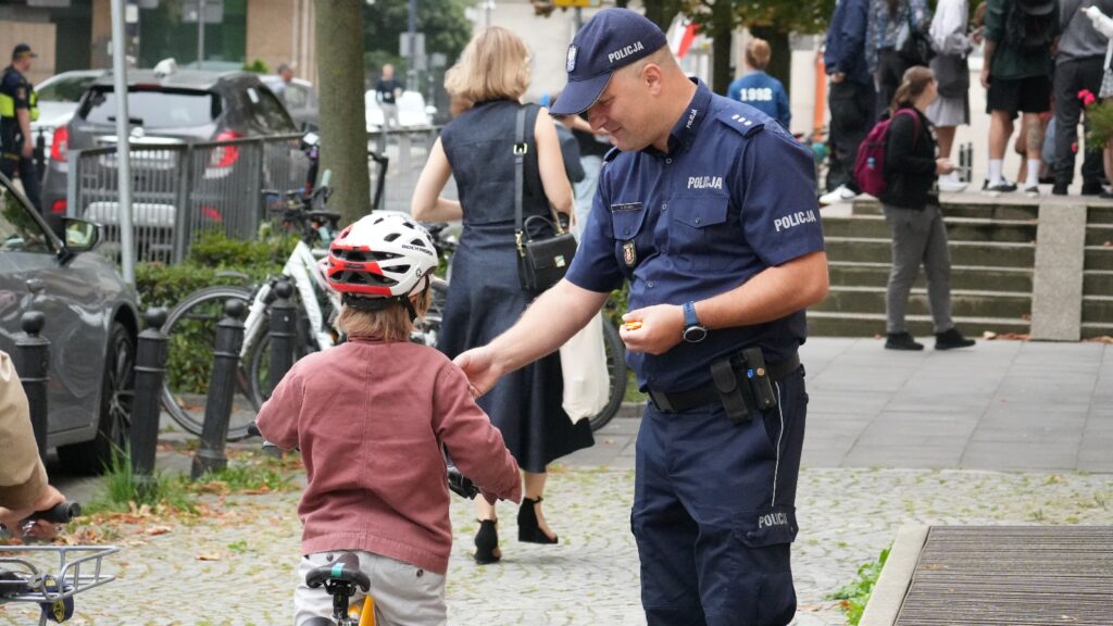 Policjant wręcza cukierka dziecku na rowerze w miejskim otoczeniu z przechodniami w tle.