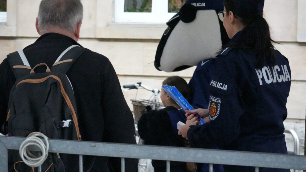 Mężczyzna z plecakiem obserwuje policjantkę wręczającą dzieciom kolorowe ulotki w miejskim otoczeniu.