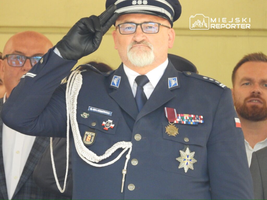 Mężczyzna w mundurze policyjnym w białych rękawiczkach oddaje honory, stojąc na tle grupy ludzi.