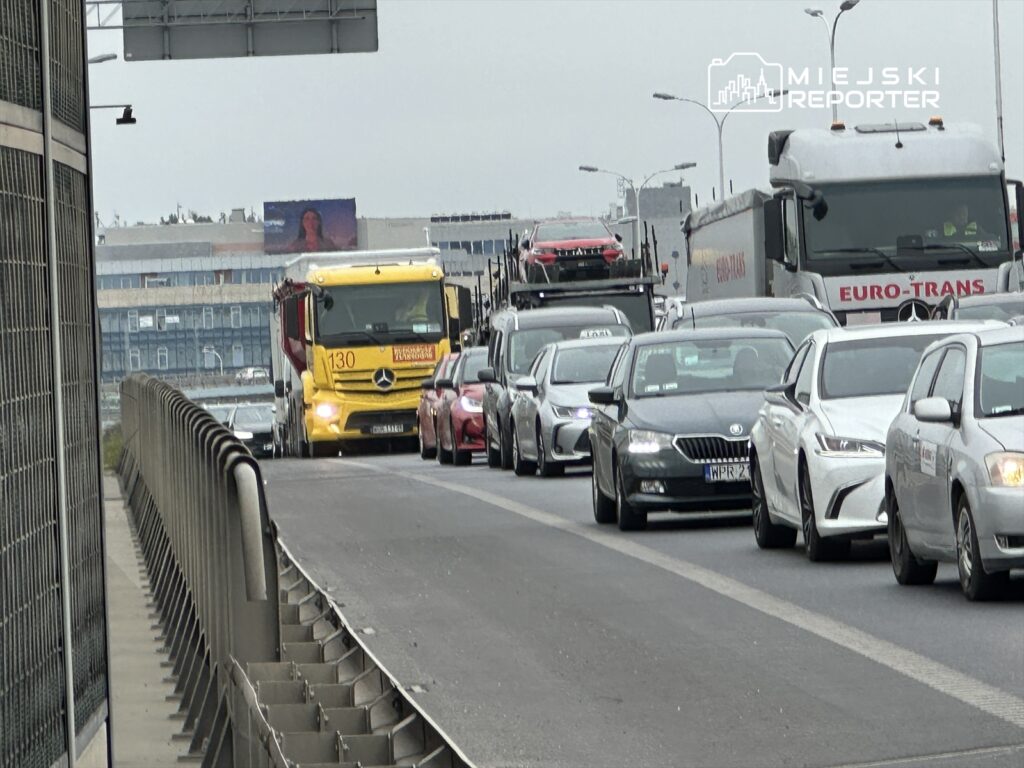 Zółty ciężarówka z napisem "130" porusza się w korku na drodze, obok innych samochodów osobowych.