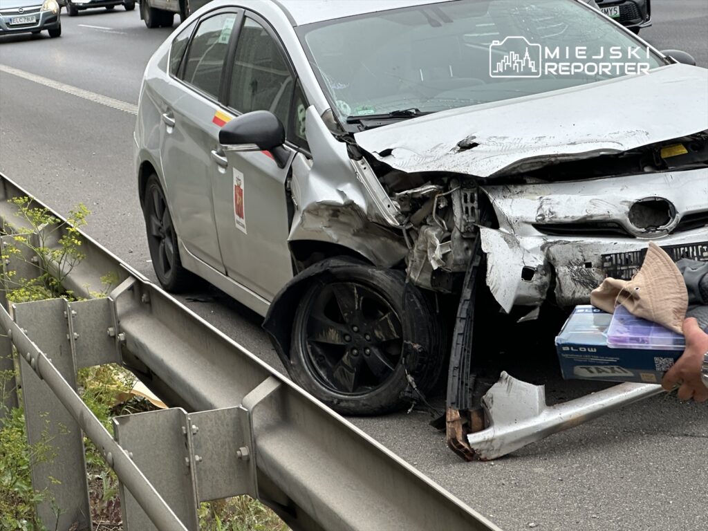Uszkodzony samochód osobowy z wgniecionym przodem stoi przy barierce na drodze, obok mężczyzna trzyma pakunek.