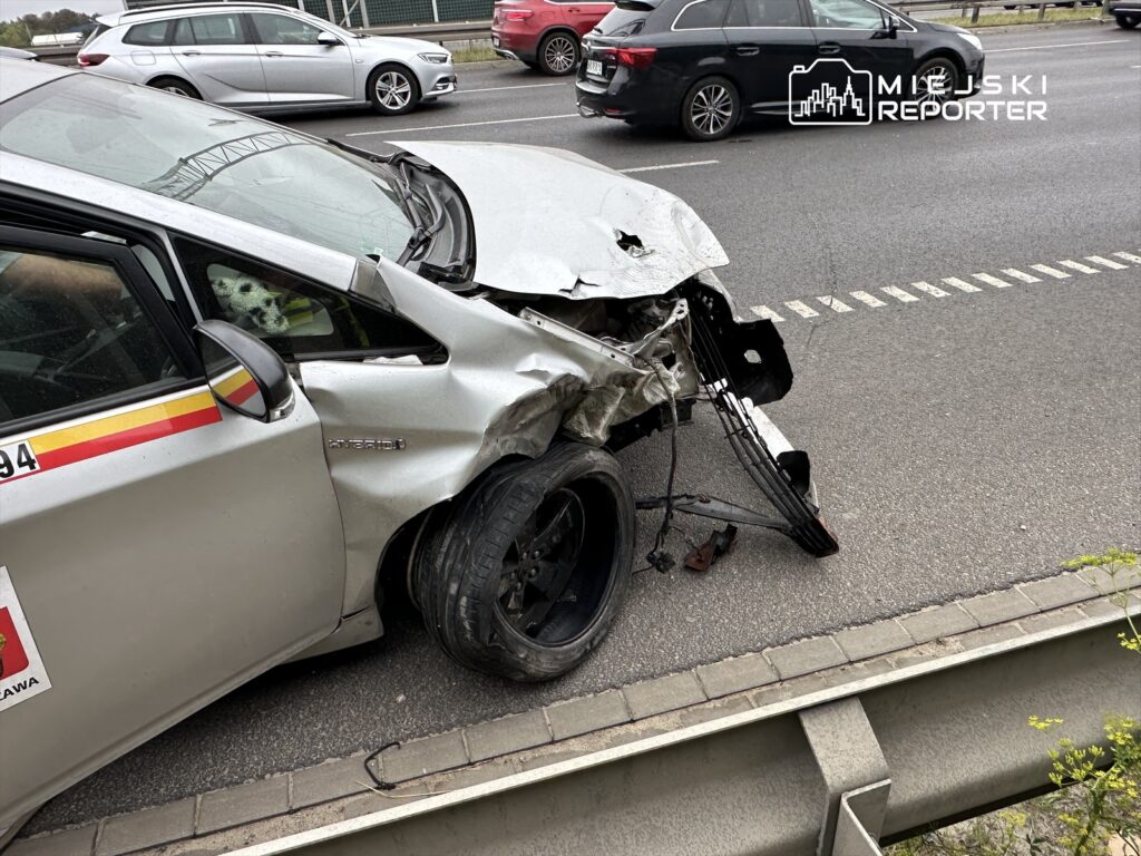 Uszkodzony samochód osobowy z wgniecioną karoserią stoi na poboczu drogi, obok innych pojazdów.