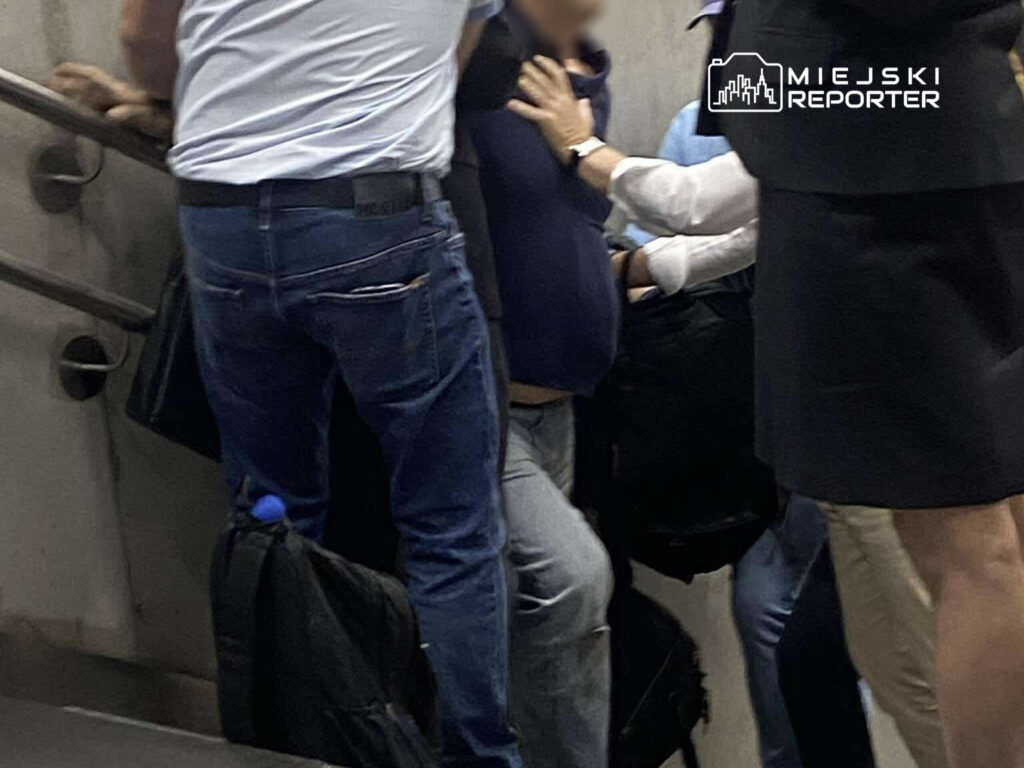 Grupa osób na schodach ruchomych, jedna osoba w niebieskim swetrze jest trzymana przez innych.