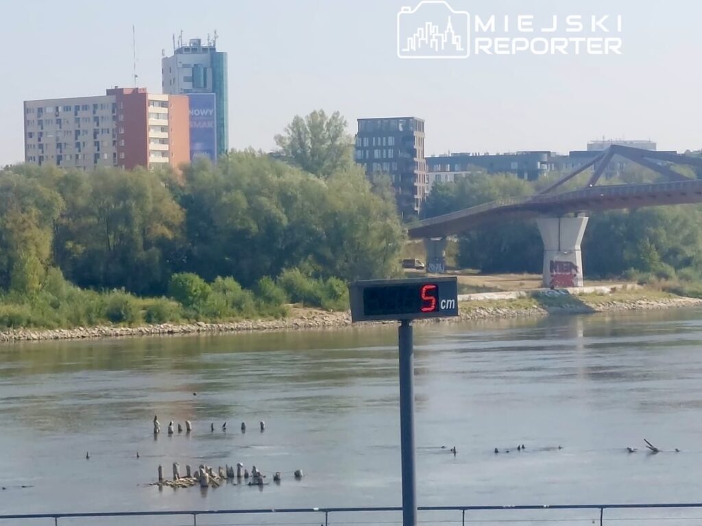 Na rzece widać słup z pomiarem poziomu wody, wskazujący 5 cm, w tle nowoczesne budynki i most.