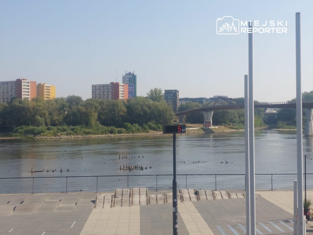 Widok na rzekę, z budynkami mieszkalnymi w tle oraz mostem nad wodą, gdzie widać pływające ptaki.