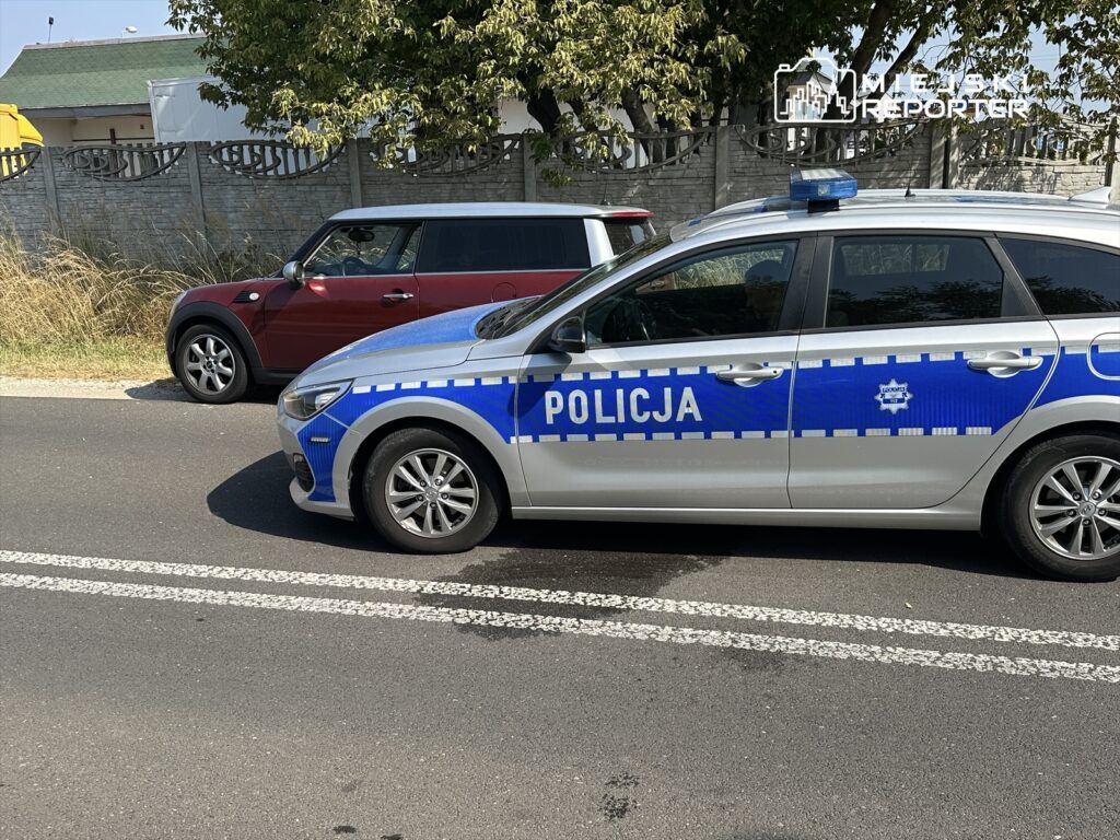 Policyjny radiowóz z niebieskimi oznaczeniami zaparkowany obok czerwonego samochodu osobowego na drodze.