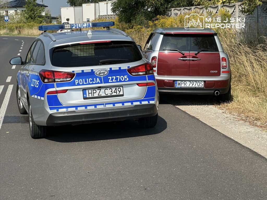 Radiowóz policyjny zatrzymany obok zaparkowanego samochodu osobowego na poboczu drogi w terenie zabudowanym.