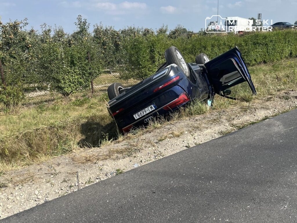 Samochód osobowy leży na dachu w rowie, obok sad z jabłoniami, w tle widoczna droga.