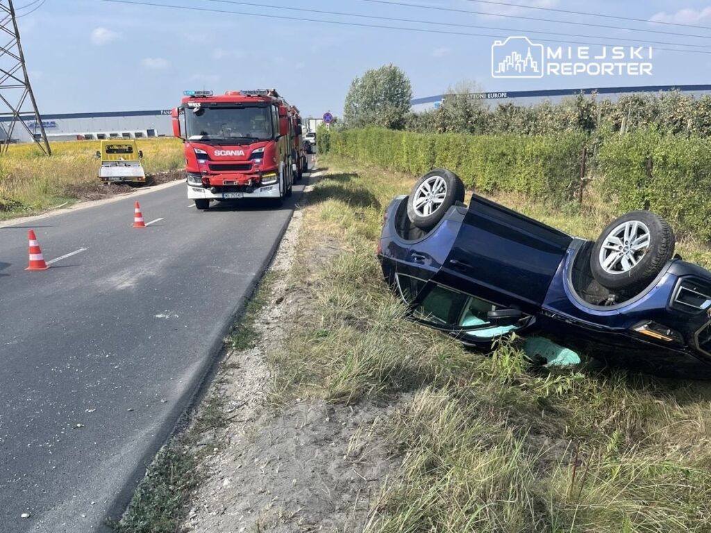 Czarny samochód przewrócony na dach przy poboczu drogi, obok stoi wóz strażacki z uruchomionymi sygnałami.