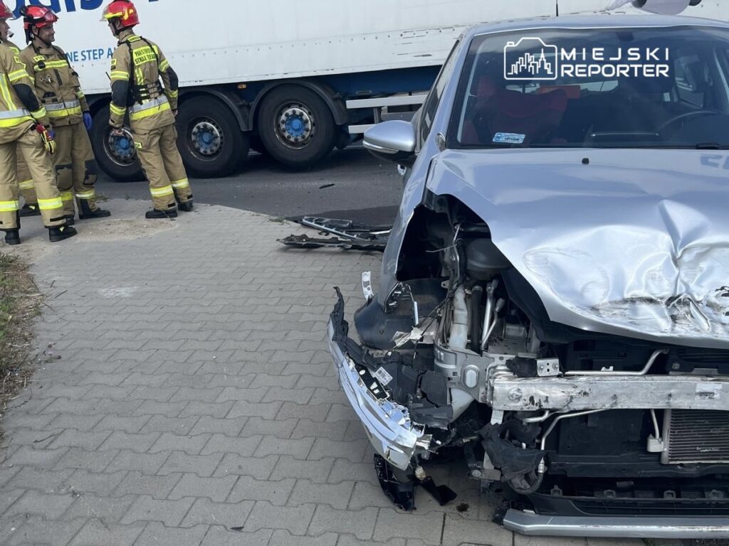 W pobliżu uszkodzonego samochodu osobowego stoją strażacy w mundurach, analizując sytuację po wypadku drogowym.