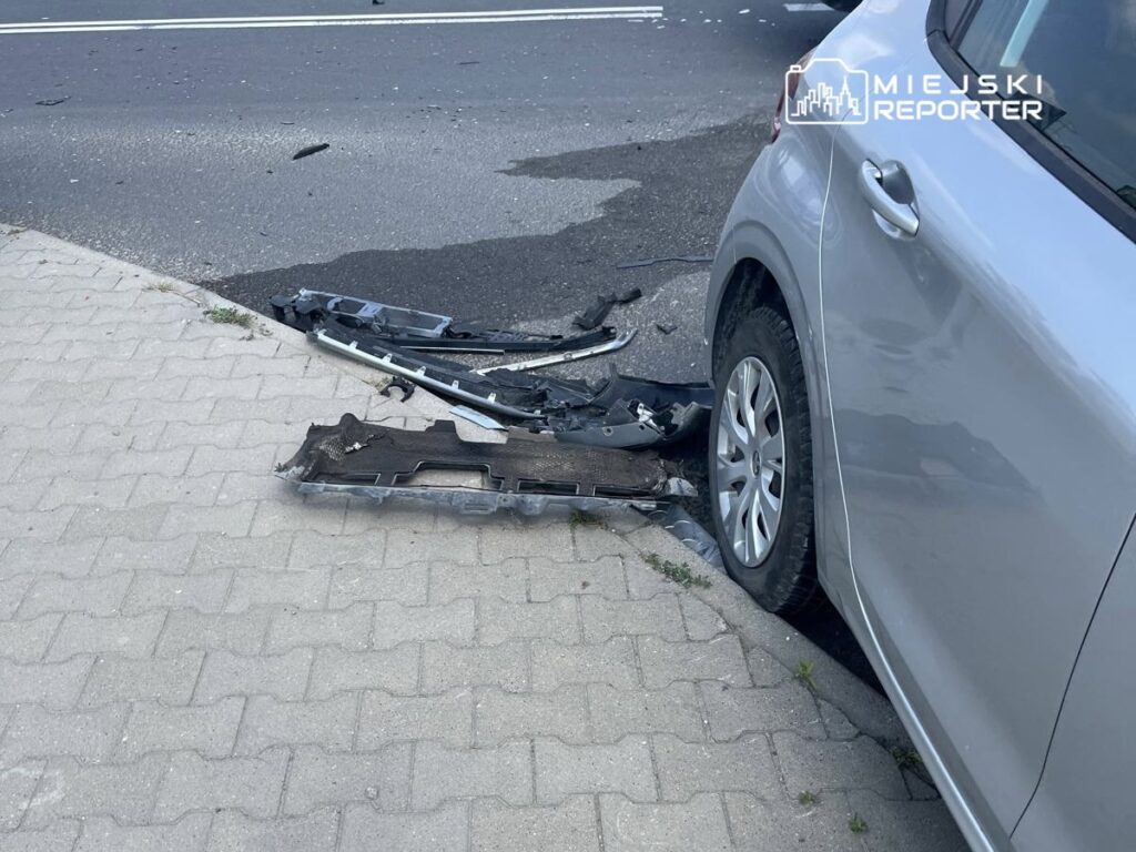 Uszkodzona karoseria samochodu osobowego leży na chodniku obok drogi, obok fragmenty zderzaka.