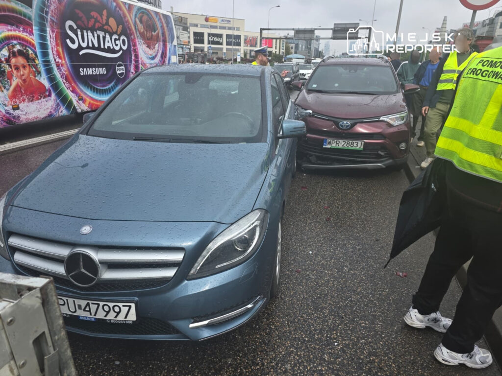Dwa uszkodzone samochody osobowe, niebieski Mercedes i bordowa Toyota, z grupą ludzi w odblaskowych kamizelkach na ruchliwej ulicy.