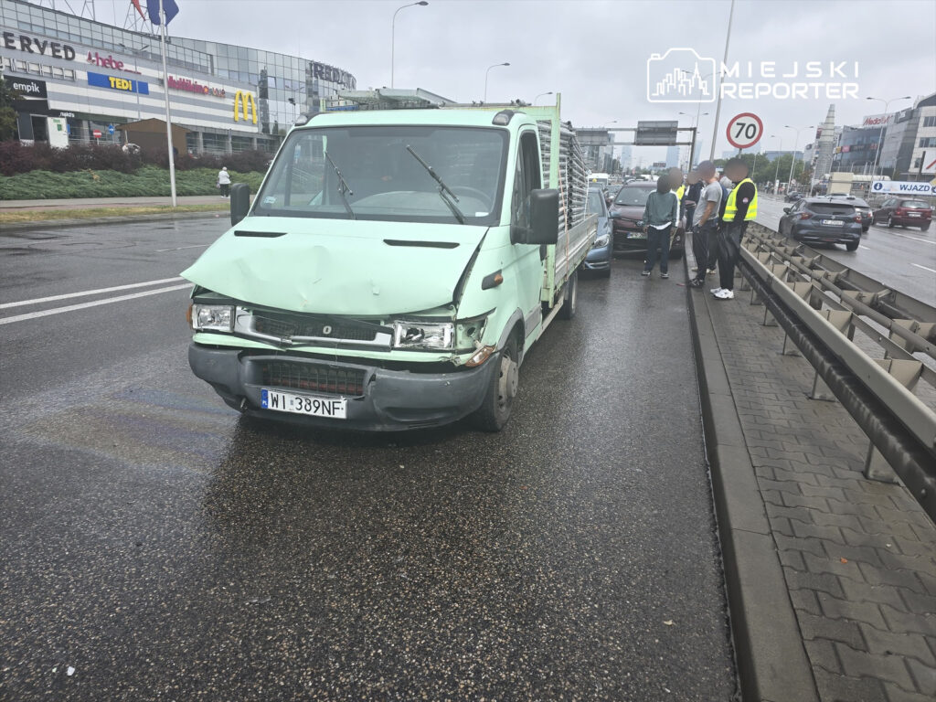 Zielony samochód dostawczy z uszkodzoną przednią częścią stoi na drodze, obok grupka osób rozmawia.