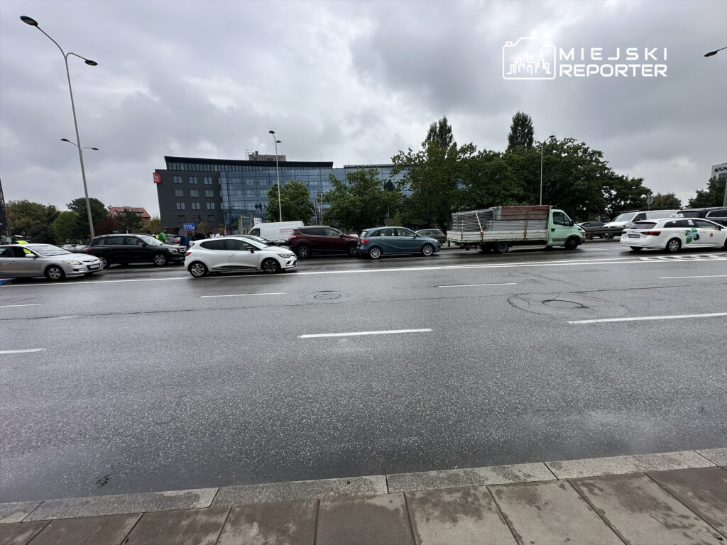 Ruch samochodowy na ulicy w deszczowy dzień, z różnymi pojazdami oraz nowoczesnym biurowcem w tle.