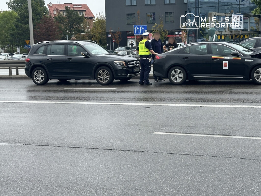 Policjant rozmawia z kierowcami dwóch samochodów na mokrej nawierzchni drogi w miejskim otoczeniu.