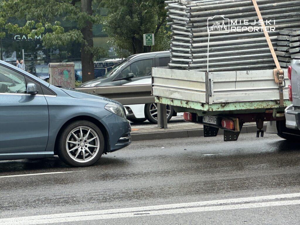 Samochód osobowy stojący obok ciężarówki z metalowymi rurami na ruchliwej ulicy w deszczowy dzień.