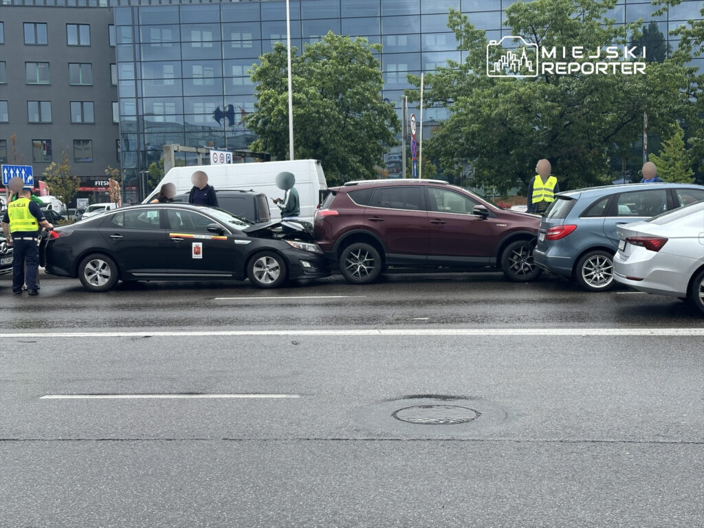 Kolizja drogowa z udziałem trzech samochodów na mokrej nawierzchni, z policjantami prowadzącymi czynności.