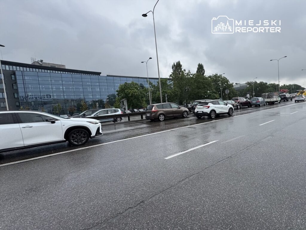 Ruch samochodowy na mokrej jezdni, w tle nowoczesny biurowiec z przeszkloną fasadą.