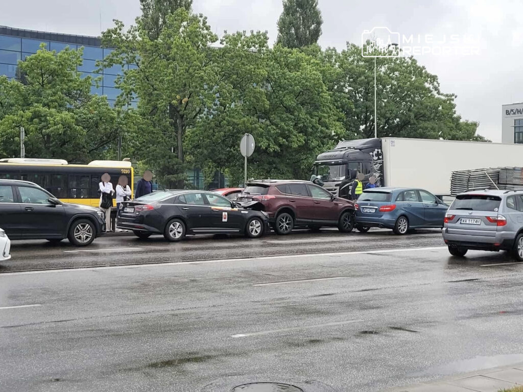 Kilkoro kierowców rozmawia obok zderzonych samochodów na mokrej ulicy w pobliżu przystanku autobusowego.