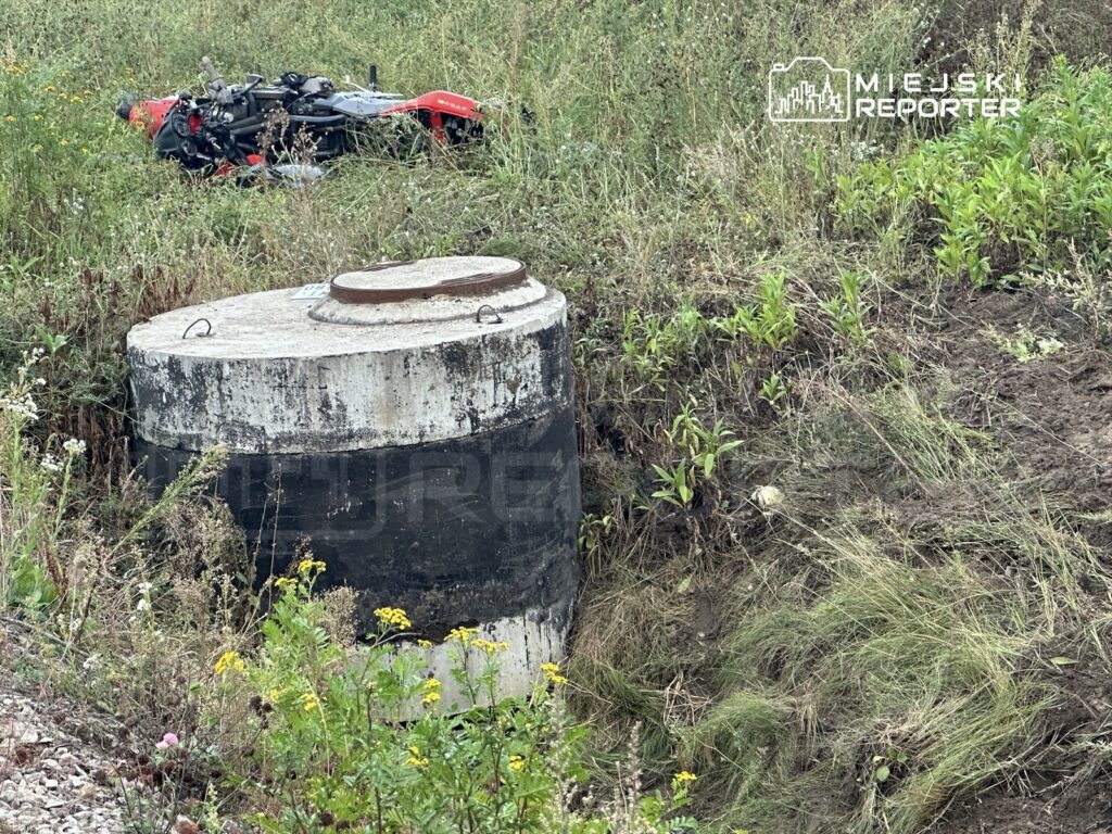 Czerwony motocykl leży na boku w wysokiej trawie obok betonowego włazu z metalową pokrywą.
