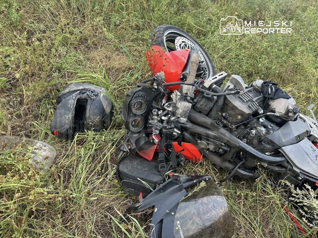Uszkodzony motocykl leży na poboczu, obok niego znajduje się zniszczony kask, a otoczenie pokryte jest wysoką trawą.