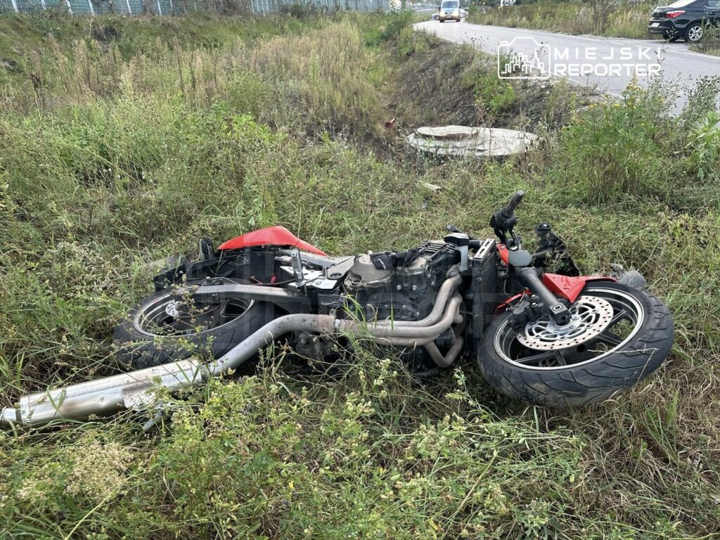 Motocykl leżący na boku w wysokiej trawie obok drogi, z widocznymi uszkodzeniami.