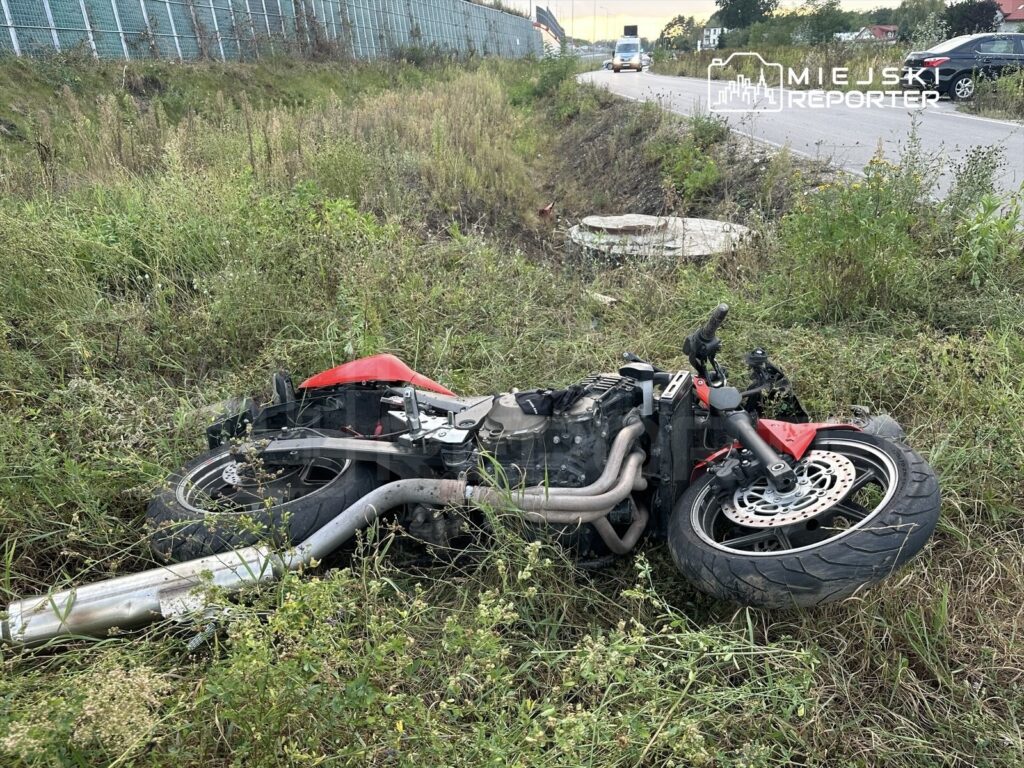 Motocykl leżący na boku w wysokiej trawie przy drodze, z widocznymi uszkodzeniami.