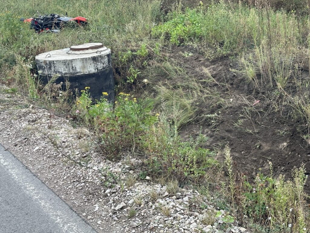 Betonowy zbiornik z metalową pokrywą w otoczeniu wysokiej trawy i krzewów, obok leży przewrócony motocykl.