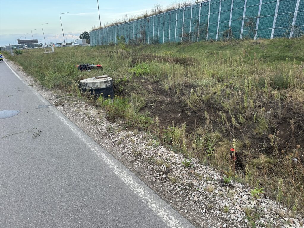 Motocykl leży na poboczu drogi, obok betonowy krąg i zarośla, w tle widoczna ogrodzona przestrzeń.