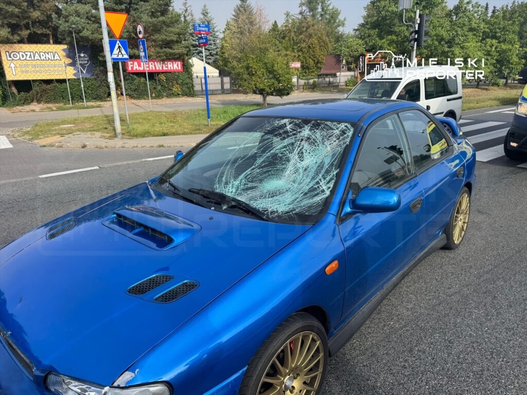 Niebieski samochód osobowy z uszkodzoną szybą przednią, stojący na drodze w miejskim otoczeniu.