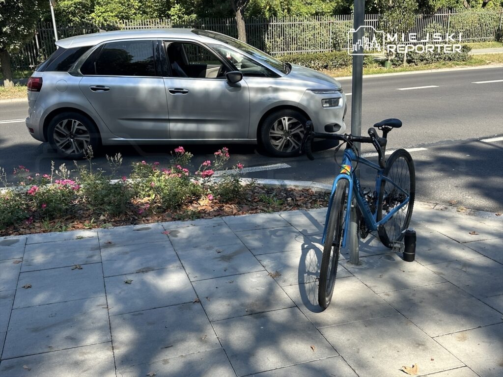 Rower na parkingu przy drodze, obok zaparkowany srebrny samochód, w tle kwitnące krzewy.