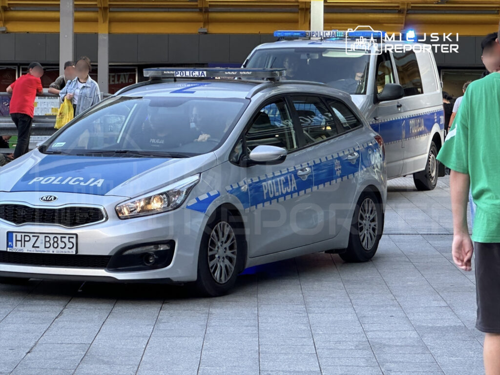 Policyjny samochód osobowy oraz furgonetka na parkingu, obok grupy ludzi rozmawiających i obserwujących sytuację.