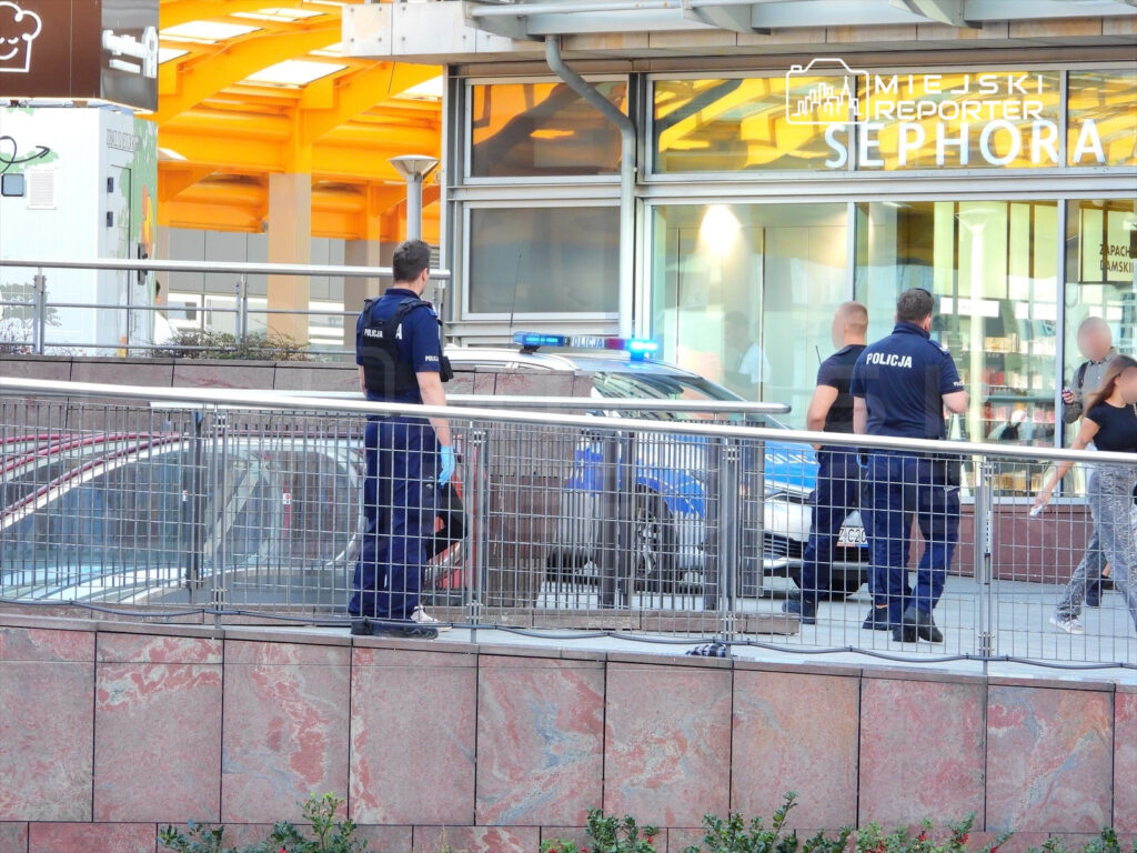 Czterech policjantów w mundurach rozmawia na chodniku przed sklepem, obok radiowozu z włączonymi sygnałami.