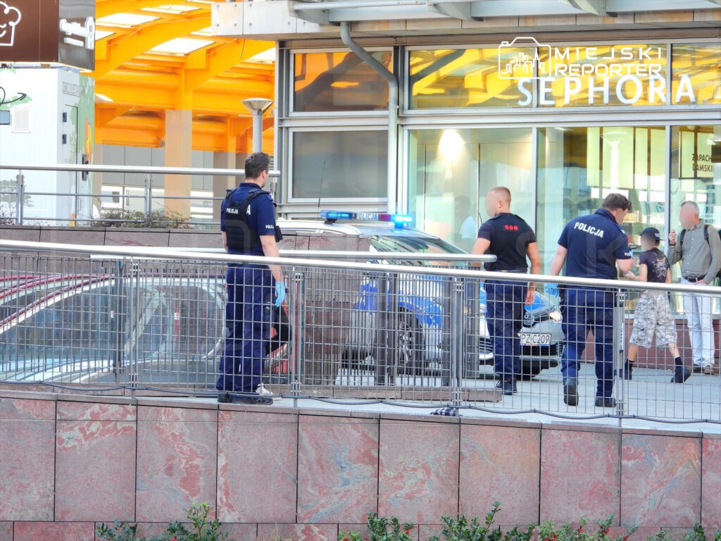 Policjanci rozmawiają z osobą na chodniku obok radiowozu przy nowoczesnym budynku handlowym.