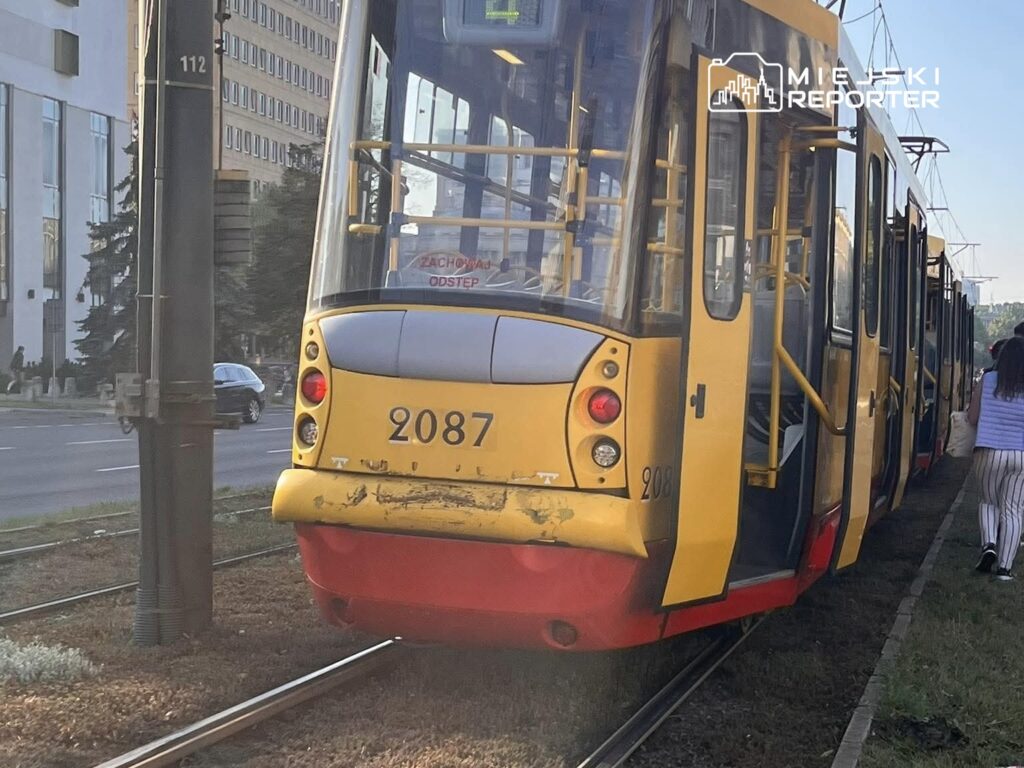 Tramwaj o numerze 2087 zatrzymany na przystanku, obok przechodzących ludzi i nowoczesnych budynków w tle.