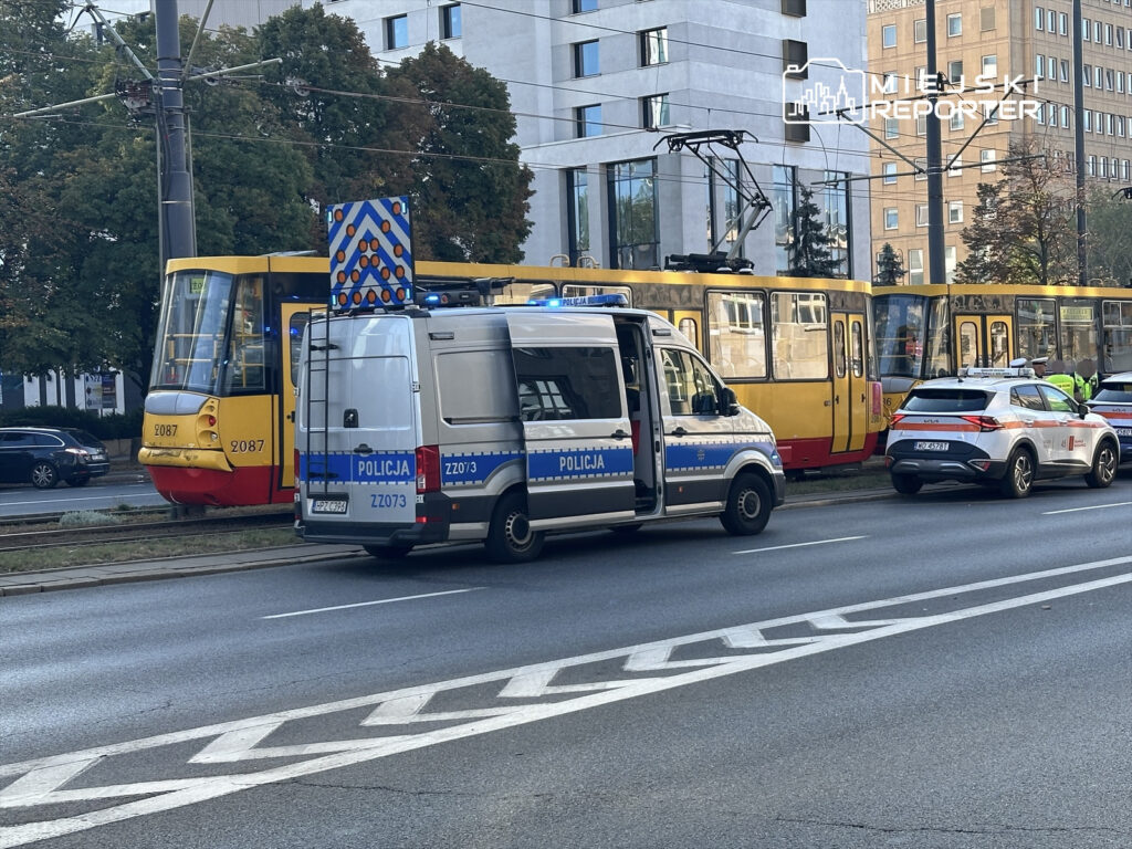 Policyjny radiowóz oraz van zaparkowane obok tramwaju na ulicy, z włączonymi sygnałami świetlnymi.
