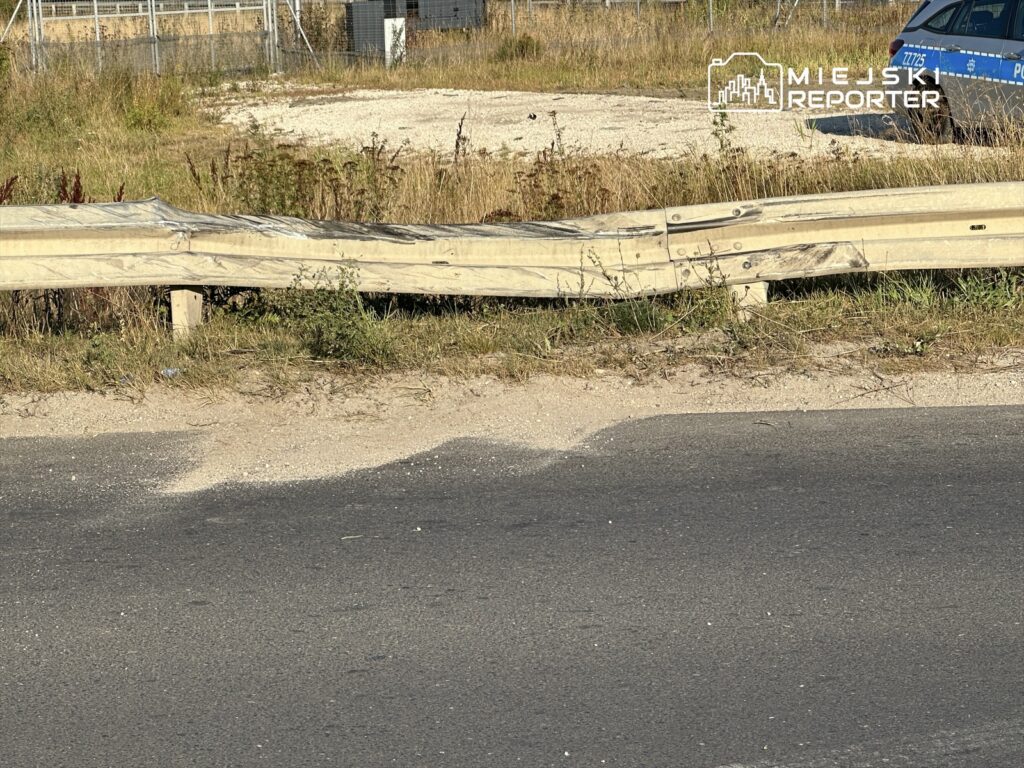 Uszkodzony fragment barierki ochronnej przy drodze, w tle widać policyjny samochód oraz zarośla.