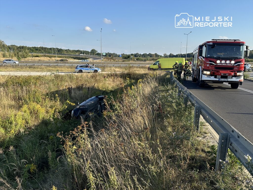 Na poboczu drogi leży przewrócone auto, obok stoją strażacy oraz woz strażacki, a w tle przejeżdżają inne pojazdy.