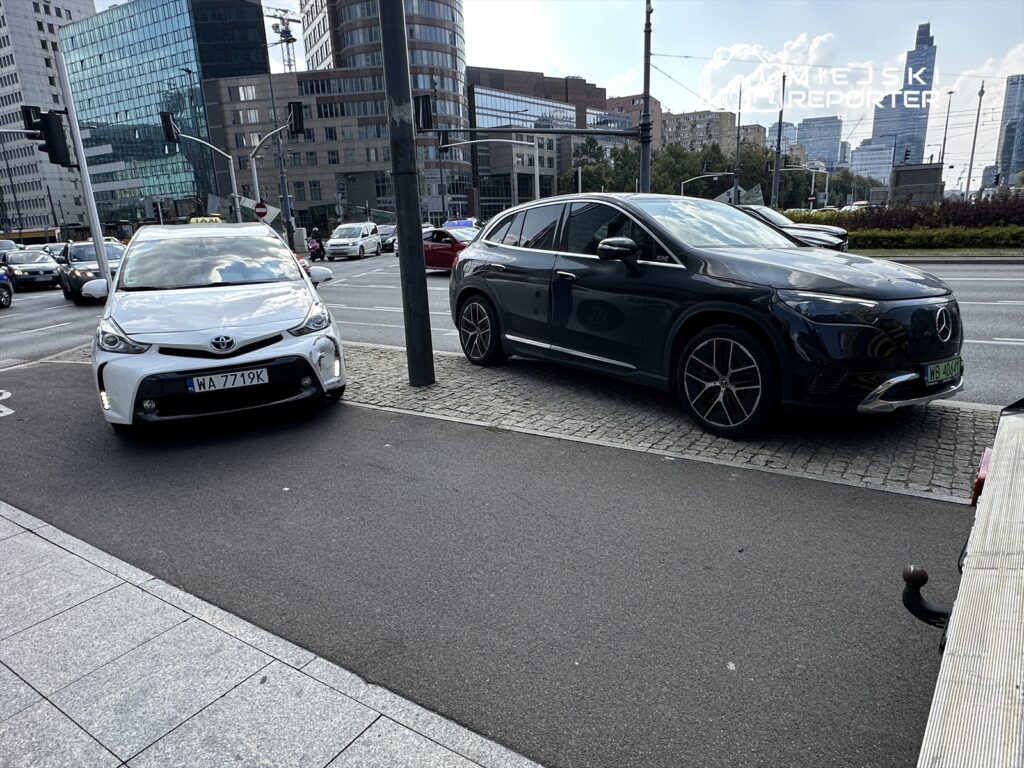 Biały samochód osobowy zaparkowany obok czarnego SUV-a na ruchliwej ulicy w miejskim otoczeniu.