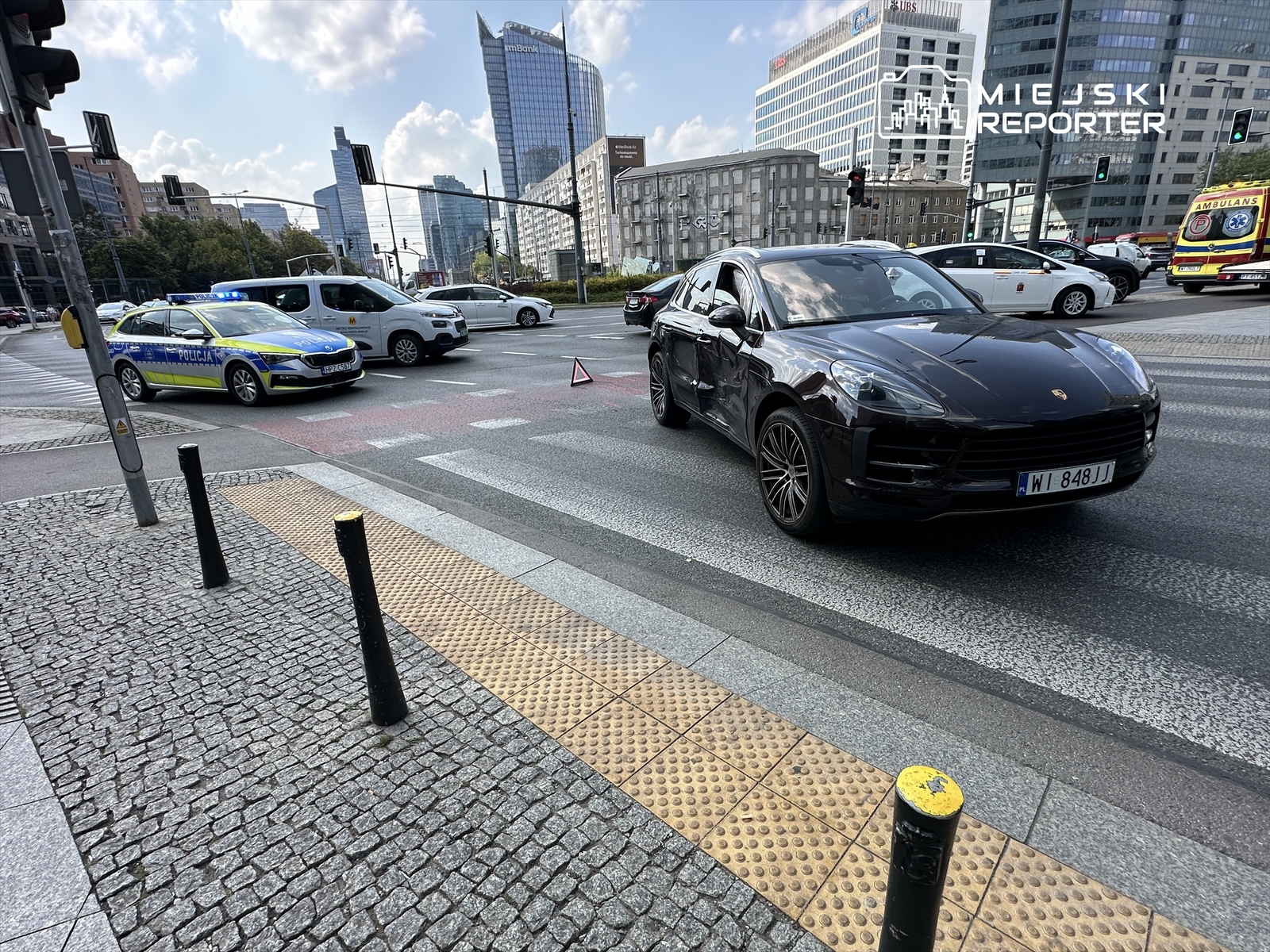 Czarny samochód sportowy Porsche zatrzymany na skrzyżowaniu, obok radiowóz policyjny z włączonymi sygnałami świetlnymi.