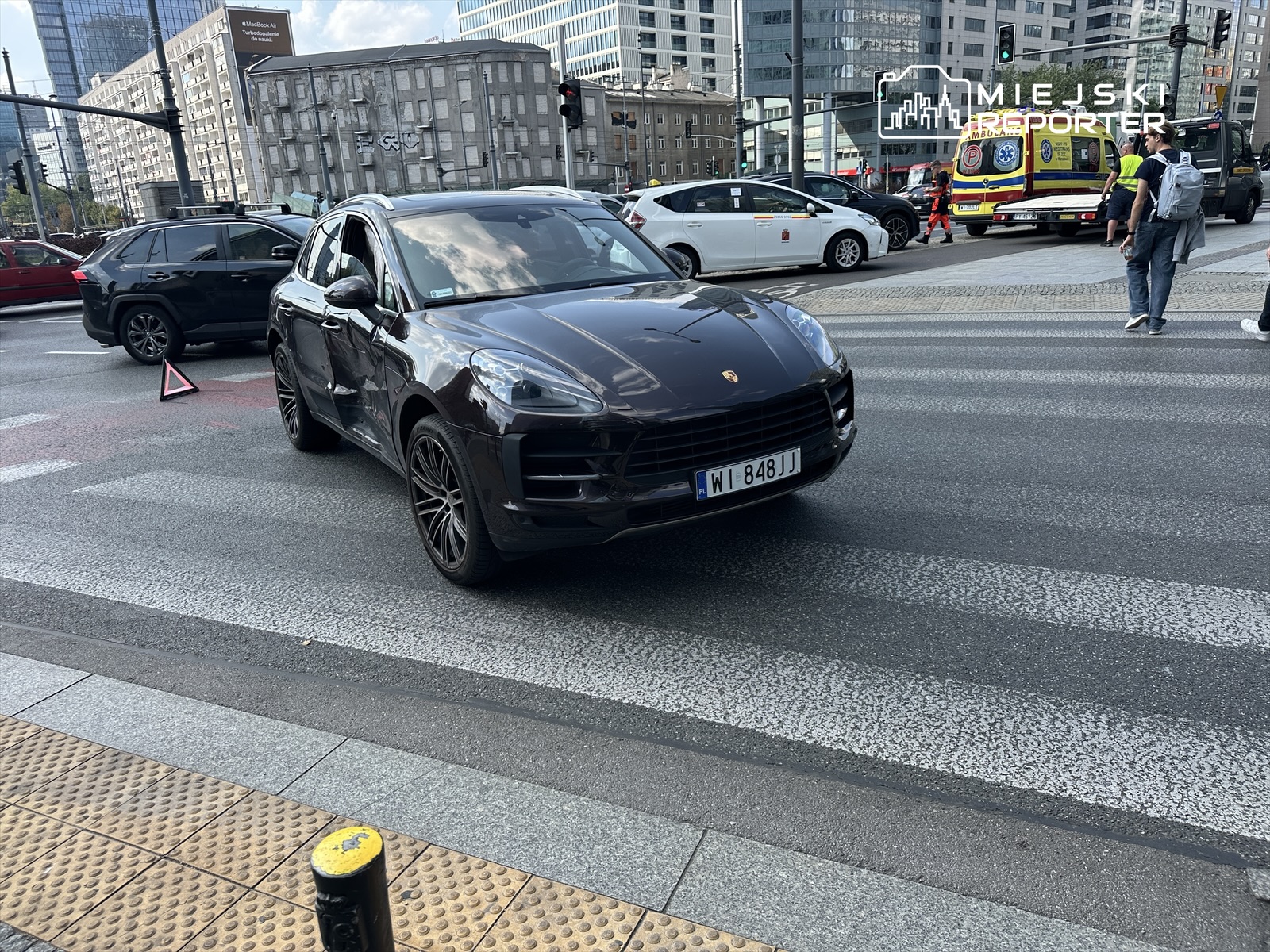 Czarny samochód SUV z uszkodzoną karoserią stoi na ulicy, obok niego widać trójkątny znak ostrzegawczy.