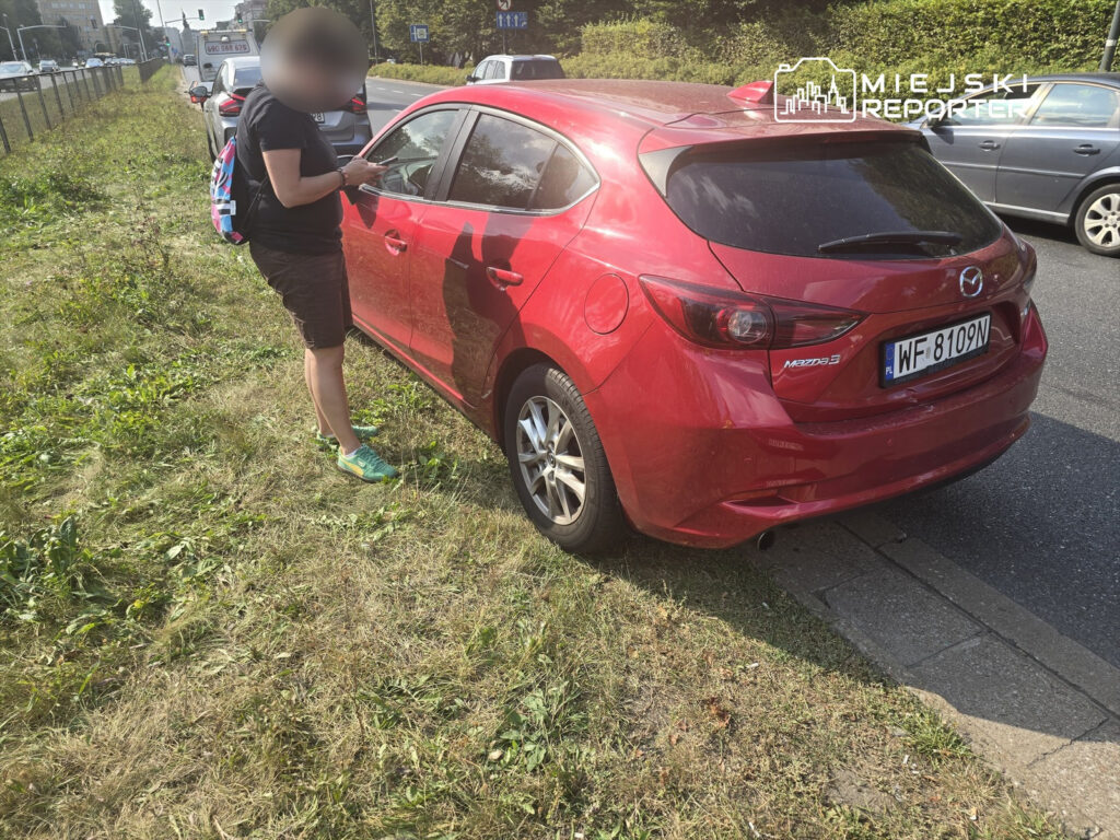 Kobieta z plecakiem korzystająca z telefonu obok czerwonego samochodu zaparkowanego na trawie przy ruchliwej drodze.
