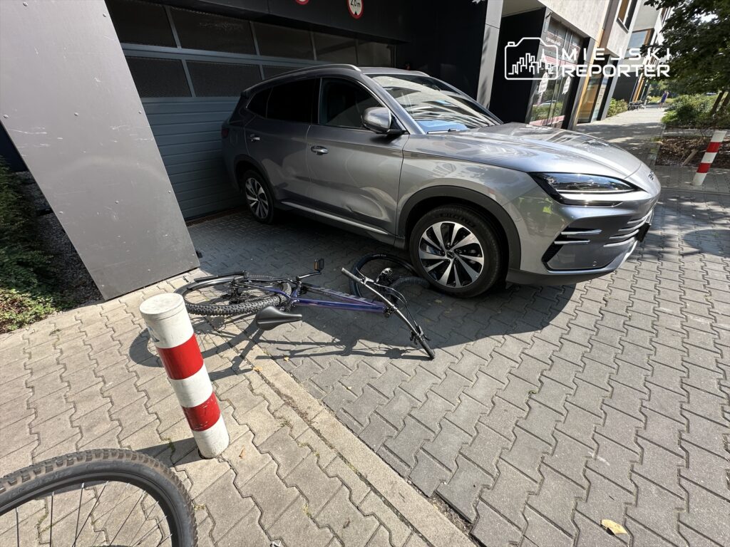 Samochód SUV w kolorze szarym stoi blisko ściany, obok leży przewrócony rower na chodniku z kostki brukowej.