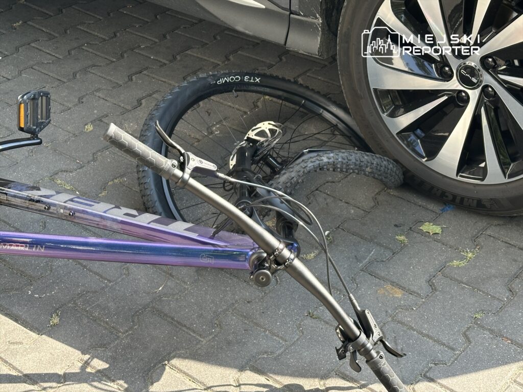Uszkodzony rower leży na chodniku obok samochodu osobowego, z jednym złamanym kołem i uchwytem kierownicy.