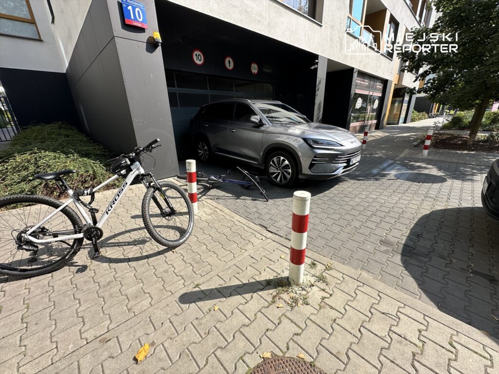 Rower górski stoi na chodniku obok nowoczesnego budynku, obok którego zaparkowany jest srebrny samochód.